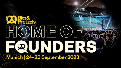 Gran audiencia observando una presentación en el escenario en el evento Bits & Pretzels, con el logotipo del evento en primer plano sobre las palabras 'Home of founders'.